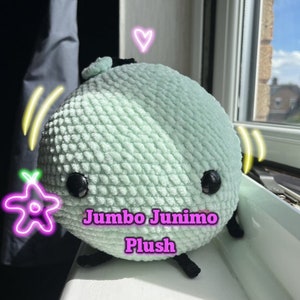 Può includere: Un grande peluche rotondo verde menta con occhi neri a bottone e piccole zampe nere. Il testo "Jumbo Junimo Plush" è visualizzato in rosa. Il peluche è appoggiato su un davanzale.