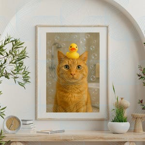 Op de afbeelding: Ingelijste print van een oranje tabbykat met een gele badeend op zijn hoofd, in een badkamer met bellen. De print wordt tentoongesteld op een houten plank met decoratieve items.