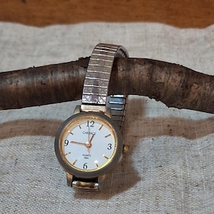 Peut inclure: Une montre-bracelet à quartz Carriage avec un cadran rond blanc, des chiffres dorés aux positions 3, 6, 9 et 12 heures, des aiguilles dorées et une fine lunette dorée. La montre possède un bracelet extensible en métal argenté.