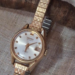May include: A gold-tone Seiko automatic wristwatch featuring a round silver dial with gold-tone hour markers, a date window at the three o'clock position, and a textured gold-tone expansion link bracelet with a repeating square pattern design.