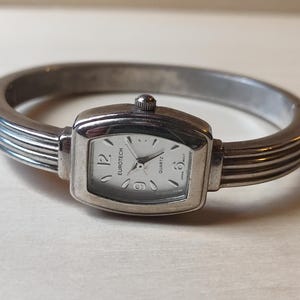 May include: Silver Eurotech quartz watch with a rectangular face and a white dial. The watch is set in a silver bangle bracelet with horizontal ridges. The watch face displays the numbers 12, 3, 6, and 9.
