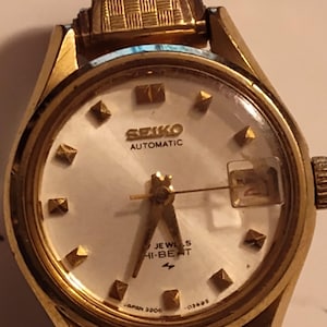 Op de afbeelding: Een goudkleurig Seiko Automatic horloge met een witte wijzerplaat en goudkleurige piramidevormige uurmarkeringen. Het horloge heeft een datumweergave en een goudkleurige band met een textuurontwerp. De wijzerplaat leest "SEIKO AUTOMATIC" en "7 JEWELS HI-BEAT".