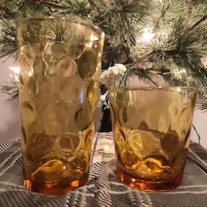 Peut inclure: Deux verres à boire ambrés avec un motif texturé en forme de bulles. L'un est un grand verre à cocktail, et l'autre est un gobelet plus court. Les verres sont posés sur une surface à carreaux, avec un sapin de Noël en arrière-plan.