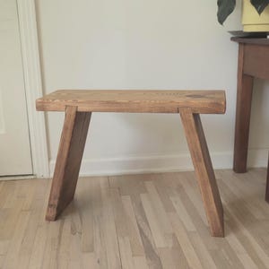 May include: A small, handcrafted wooden stool with a rectangular seat and angled legs. The stool is made of light brown wood and has a simple, rustic design. It is approximately 12 inches tall and 18 inches wide.