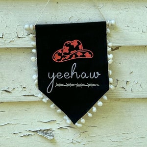 Pode incluir: Bandeira de feltro preto com acabamento em pompons brancos. A bandeira apresenta um chapéu de caubói vermelho e preto com a palavra "yeehaw" bordada em branco.