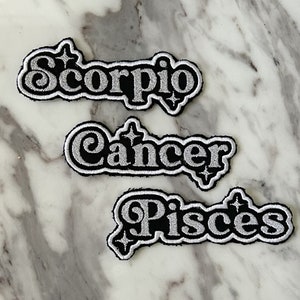 May include: Three iron-on patches with silver glitter lettering on a black background. The patches read "Scorpio", "Cancer", and "Pisces".