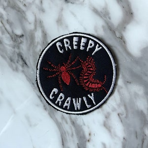 May include: A black and white embroidered patch with the words "Creepy Crawly" in white text. The patch features a red and white spider and a red and white centipede.