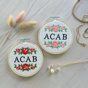 Puede incluir: Dos aros bordados con el texto "ACAB" rodeados de flores rosas y rojas. Los aros están sobre una superficie de madera blanca.