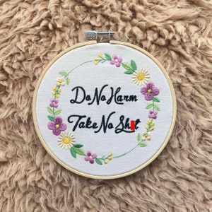 以下が含まれることがあります： 白い背景と花輪のデザインが施された刺繍のフープアート。"Do No Harm Take No Shit"というテキストが黒い筆記体で刺繍されています。