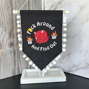 Könnte beinhalten: Schwarzes Wimpelbanner mit weißem Pomponbesatz und dem Text "F*ck Around And Find Out". Das Banner zeigt einen roten Teufel mit Hörnern und Flammen. Das Banner wird auf einem weißen Ständer angezeigt.