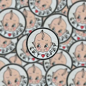 May include: A close-up of several iron-on patches featuring a cartoon baby with a teardrop and the text "Cry Baby".