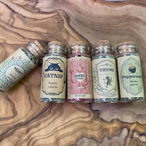 May include: Five small glass jars with cork stoppers, each containing dried herbs or petals. The labels feature decorative borders and text: Mint, Catnip with a cat illustration, Rose Petals, Thyme with a lion illustration, and Peppermint with a cauldron.