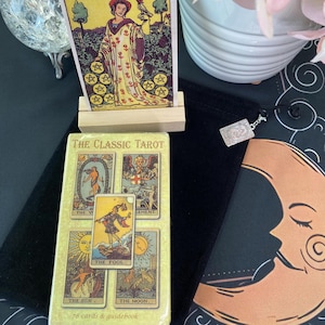 May include: A deck of The Classic Tarot cards in a box featuring illustrations of The World, Judgement, The Sun, The Moon, and The Fool. A Nine of Pentacles card stands on a wooden holder, and a small silver-toned charm hangs from a black velvet bag.