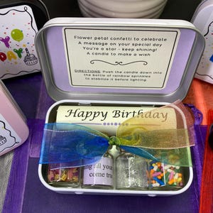 May include: Open metal tin with a Happy Birthday message, small glass bottles filled with confetti, glitter, and sprinkles, and a ribbon. The tin's lid has a message about celebrating a special day. The tin is open, revealing its contents.
