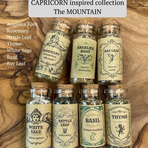 May include: Seven clear glass apothecary jars with cork stoppers, each containing dried herbs labeled Rosemary, Angelica Root, Bay Leaf, White Sage, Nettle Leaf, Basil, and Thyme. The text reads CAPRICORN inspired collection The MOUNTAIN.