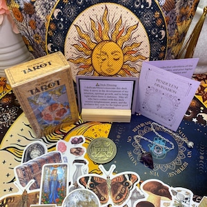 May include: A collection of tarot cards, stickers, and a pendulum. The tarot card box is tan with the words "The Original Tarot" printed on it. Stickers include a butterfly, a coin, and a mushroom. A purple pendulum is also visible.