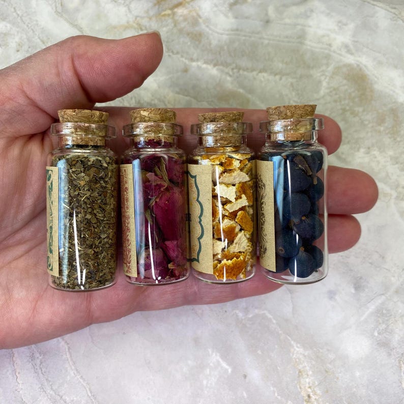 May include: Four small clear glass vials with cork stoppers, each containing different dried botanical ingredients: green herbs, pink rose petals, orange citrus peel, and dark blue juniper berries, all featuring tan paper labels.