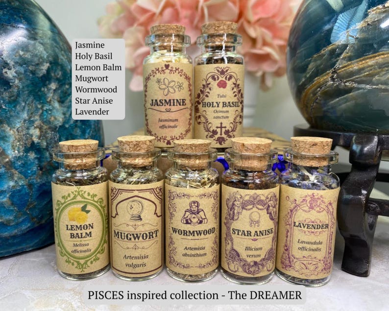 May include: Seven small glass jars with cork stoppers, each containing dried herbs labeled Jasmine, Holy Basil, Lemon Balm, Mugwort, Wormwood, Star Anise, and Lavender. The labels feature decorative borders and botanical names in a tan paper finish.
