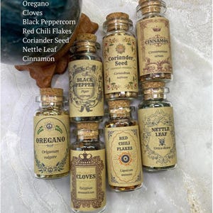 May include: A collection of seven clear glass spice jars with cork stoppers, each featuring a tan label with decorative borders and text including ARIES inspired collection - FLAME of the WARRIOR, Oregano, Cloves, Black Pepper, Coriander Seed, Red Chili Flakes, Nettle Leaf, and Cinnamon.