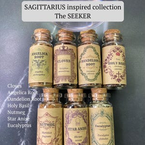 May include: Seven small glass jars with cork stoppers, each containing dried herbs or spices with labels reading Angelica Root, Cloves, Dandelion Root, Holy Basil, Nutmeg, Star Anise, and Eucalyptus. A sign above reads SAGITTARIUS inspired collection The SEEKER.