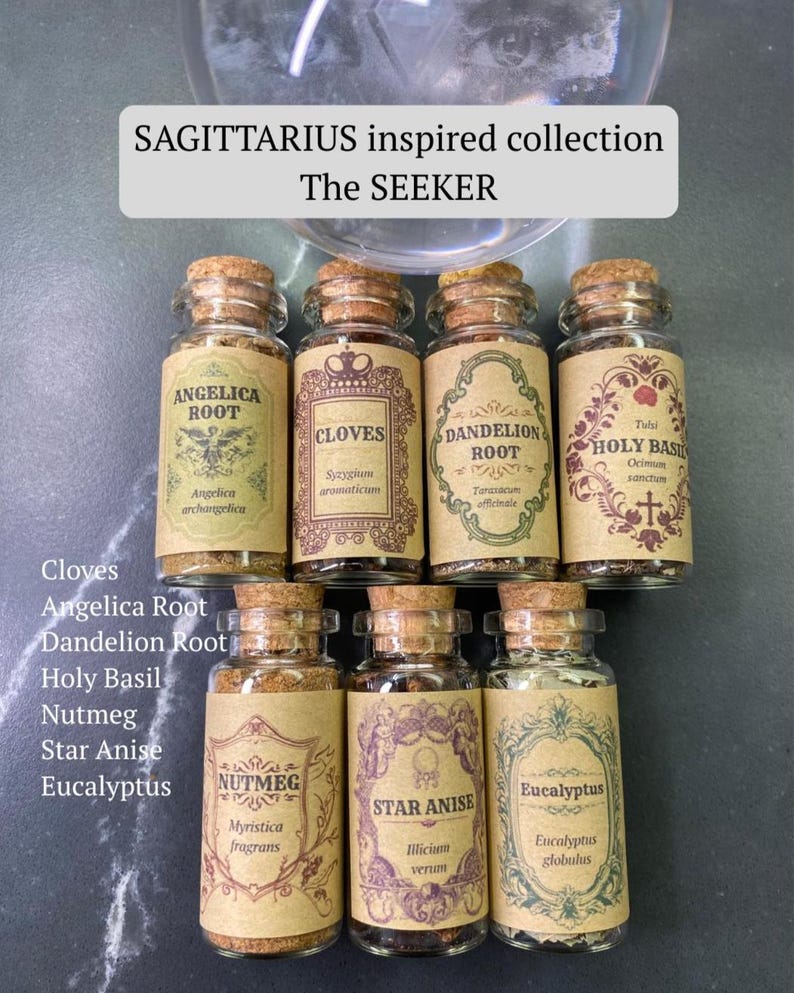May include: Seven small glass jars with cork stoppers, each containing dried herbs or spices with labels reading Angelica Root, Cloves, Dandelion Root, Holy Basil, Nutmeg, Star Anise, and Eucalyptus. A sign above reads SAGITTARIUS inspired collection The SEEKER.