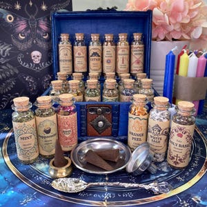 May include: A collection of small glass jars with cork stoppers, each containing dried herbs and labeled with names like Mint, Peppermint, Rose Petals, Orange Peel, White Sage, and Holy Basil. Includes a metal spoon, funnel, and incense cones.