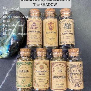 May include: A collection of seven small glass jars with cork stoppers, each containing dried herbs or seeds. Labels feature text: Wormwood, Cilantro, Black Cumin Seed, Basil, Coriander Seed, Mugwort, and Black Pepper. A large, iridescent stone sits to the left.