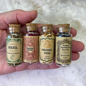 May include: Four small clear glass vials with cork stoppers, each containing dried botanicals. The labels feature ornate borders and text: Basil, Rose Petals, Orange Peel, and Indian Hawthorn Berries, along with their botanical names.