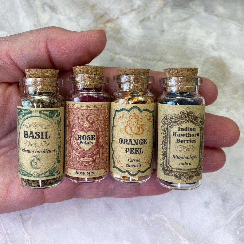 May include: Four small clear glass vials with cork stoppers, each containing dried botanicals. The labels feature ornate borders and text: Basil, Rose Petals, Orange Peel, and Indian Hawthorn Berries, along with their botanical names.