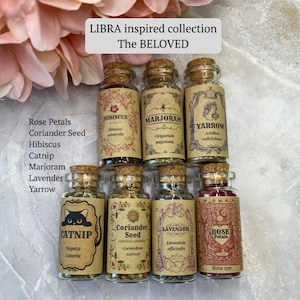 May include: A collection of eight small glass jars with cork stoppers, each containing dried herbs or flowers. Labels feature text including LIBRA inspired collection, The BELOVED, Hibiscus, Marjoram, Yarrow, Catnip, Coriander Seed, Lavender, and Rose Petals.