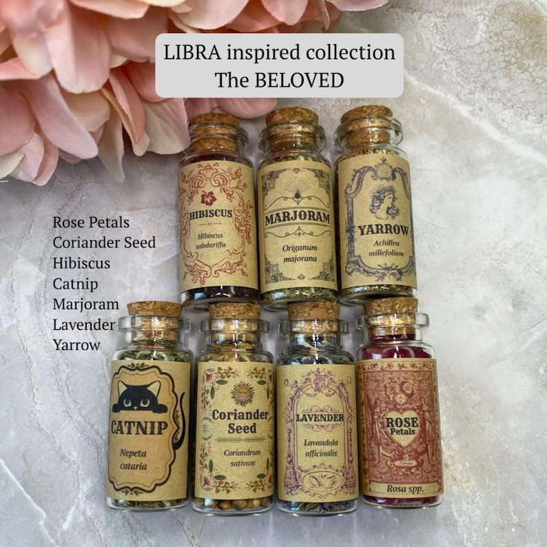 May include: A collection of eight small glass jars with cork stoppers, each containing dried herbs or flowers. Labels feature text including LIBRA inspired collection, The BELOVED, Hibiscus, Marjoram, Yarrow, Catnip, Coriander Seed, Lavender, and Rose Petals.