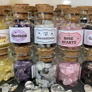 Könnte beinhalten: Acht kleine Glasflaschen gefüllt mit verschiedenen Kristallsorten. Die Flaschen sind mit den Namen der Kristalle beschriftet: Citrin, Amethyst, Mondstein, Rosenquarz und Granat.