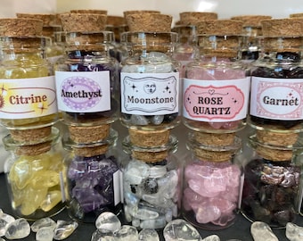 Könnte beinhalten: Acht kleine Glasflaschen gefüllt mit verschiedenen Kristallsorten. Die Flaschen sind mit den Namen der Kristalle beschriftet: Citrin, Amethyst, Mondstein, Rosenquarz und Granat.