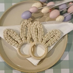 May include: Two beige crocheted bunny ear napkin rings with wooden rings. The rings are on a white napkin on a beige plate.