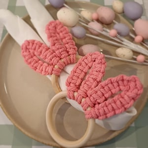 May include: Two pink macrame bunny ear napkin rings on a beige plate with white napkins. Decorative Easter eggs in pastel colours are in the background. The napkin rings have wooden rings and are made of a woven pink material.