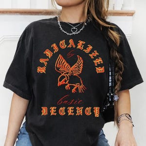 May include: Black t-shirt with the words "RADICALIZED by BASIC DECENCY" in orange lettering. The shirt features an orange eagle graphic. The model is wearing blue jeans and fishnet stockings.