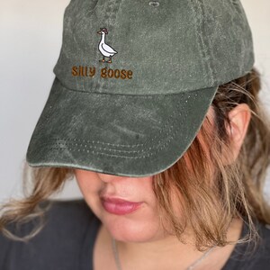 K&ouml;nnte beinhalten: Olivgr&uuml;ne Baseballkappe mit gebogenem Schirm. Die Kappe hat ein gesticktes Design einer Gans mit Hut, mit den Worten "Silly goose" in brauner Schrift.