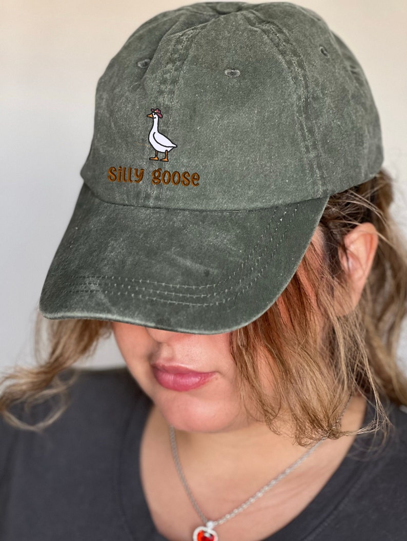 K&ouml;nnte beinhalten: Olivgr&uuml;ne Baseballkappe mit gebogenem Schirm. Die Kappe hat ein gesticktes Design einer Gans mit Hut, mit den Worten "Silly goose" in brauner Schrift.