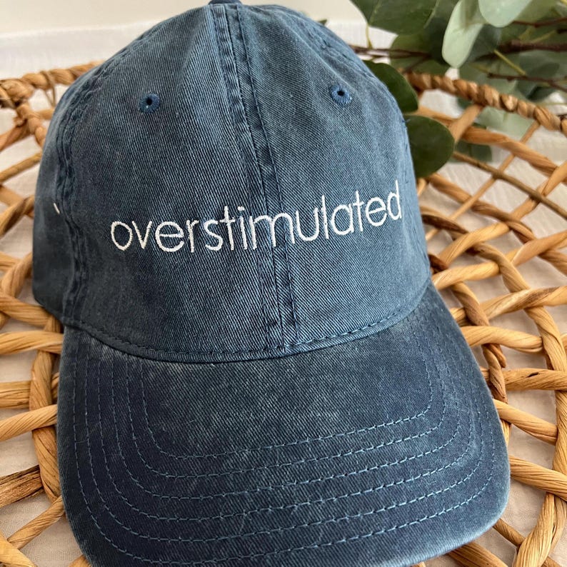 May include: A dark blue baseball cap with the word "overstimulated" embroidered in white on the front.