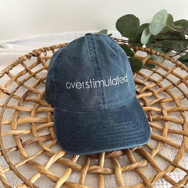 May include: A dark blue baseball cap with the word "overstimulated" embroidered in white on the front.