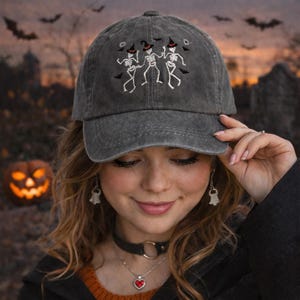 May include: Gray baseball cap with an embroidered design of three dancing skeletons wearing witch hats. The cap is a classic six-panel style with a curved brim. The design is in white thread, creating a spooky Halloween theme.