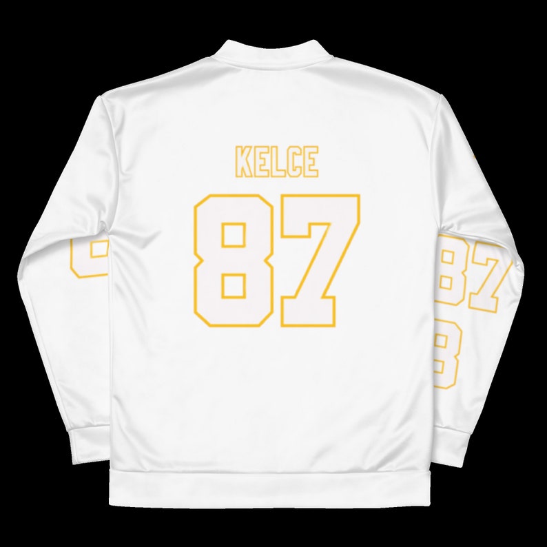 Puede incluir: Chaqueta blanca de manga larga con cremallera, con el nombre "KELCE" y el n&uacute;mero "87" impresos en amarillo en la espalda. El n&uacute;mero "87" tambi&eacute;n est&aacute; impreso en las mangas.