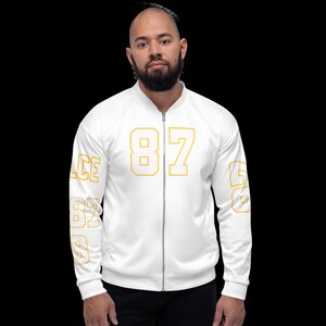 Puede incluir: Chaqueta bomber blanca con un contorno dorado del n&uacute;mero 87 en la parte delantera y contornos dorados de los n&uacute;meros 87 y 6 en las mangas.