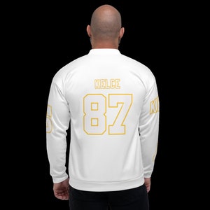 Puede incluir: Chaqueta bomber blanca con letras doradas en la espalda. Las letras dicen "KELCE 87".
