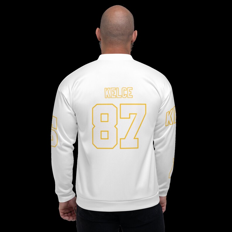 Puede incluir: Chaqueta bomber blanca con letras doradas en la espalda. Las letras dicen "KELCE 87".