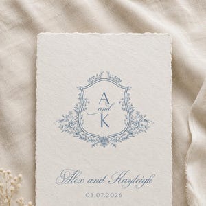 Puede incluir: Una invitación crema con un escudo floral azul con las letras "A" y "K". Los nombres "Alex and Kayleigh" y la fecha "03.07.2026" están impresos debajo. El papel tiene un borde texturizado y rasgado.