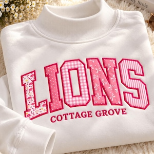 May include: A white mock neck sweatshirt featuring the word LIONS in large, pink-bordered block letters with varied patterns including floral, polka dot, gingham, and stripes. The text COTTAGE GROVE is embroidered in pink below the main word.