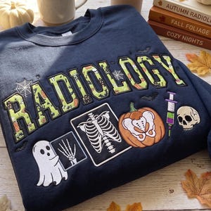 May include: A navy blue crewneck sweatshirt featuring the embroidered word RADIOLOGY in green patterned lettering. Below, embroidered graphics include a ghost holding an X-ray of a hand, a rib cage X-ray, a pumpkin, a syringe, and a human skull.
