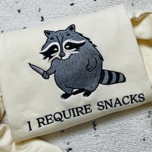 May include: Cream-colored sweatshirt featuring an embroidered raccoon holding a knife. The raccoon is gray and white, with black accents. The text "I REQUIRE SNACKS" is embroidered below the raccoon in black.