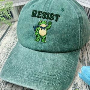 May include: A green baseball cap with the word "RESIST" embroidered above a cartoon frog wearing a blue scarf. The frog is waving its right hand. The cap has a curved brim and a button on top.